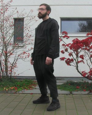 Ein Mann vor einer Hauswand in einem komplett schwarzen gemütlichen Techwear Outfit bestehend aus Uniqlo Shirt, Uniqlo Jacke und idle/ido Techwear Hose mit schwarzen Sneakern