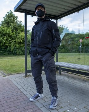 Techwear.Germany aka Janek vor einer Bushaltestelle in einem schwarz-grauen Techwear Outfit bestehend aus North Face Cap, Maske, Orbit Gear W-110j Techwear Jacke, Enfin Levé Ameztu Stotz Etaproof Techwear Hose und 11byBBS x Salomon Bamba 2 Low Techwear Sneaker