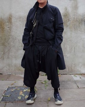 A man in an all-black techwear outfit in front of a concrete wall consisting of an ACRONYM J1AL-GT techwear coat, HAMCUS techwear pants and Nike ACG Mountain Fly techwear sneakers