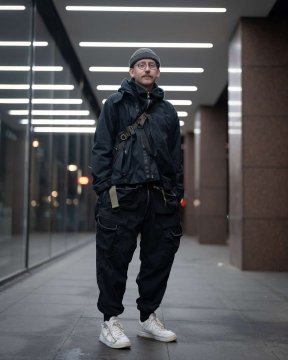 A next to a futuristic building in complete techwear outfit consisting of an ACRONYM techwear jacket, an ACRONYM techwear bag, ACRONYM P38-E pants and ACRONYM x Nike Lunar Force techwear sneaker